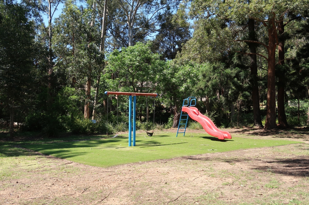 Tiny playground with two swings and a slide, surrounded by trees and carpeted with artificial grass.